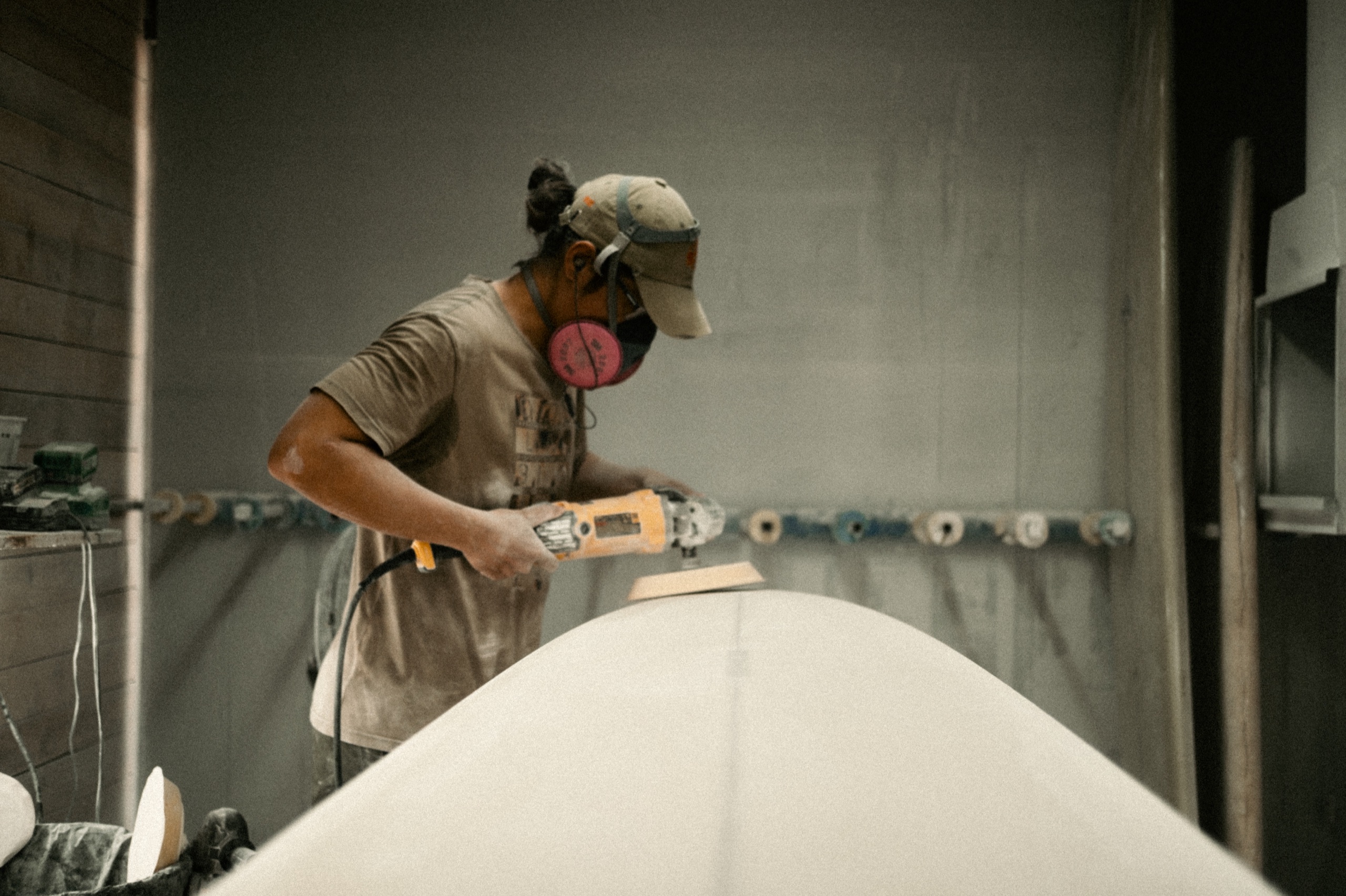 A masked board builder uses a power sander on a white surfboard.