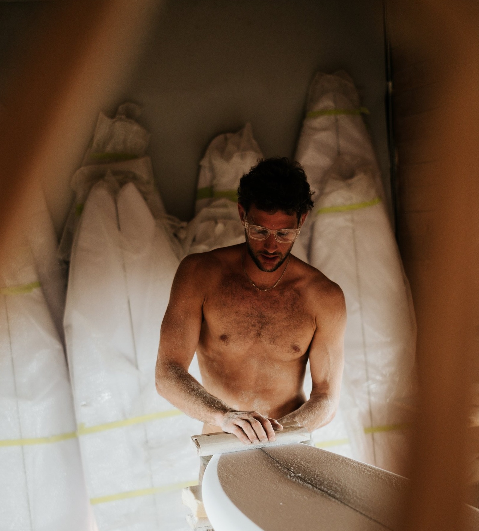 A surfboard builder sands a board in front of a rack of finished boards.