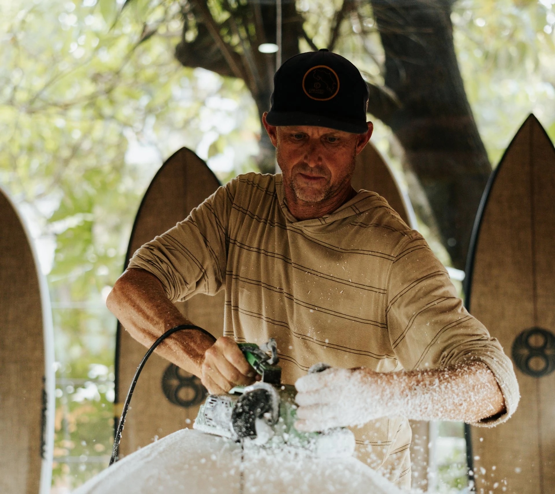 A surfboard shaper works with a power tool as foam dust flies.