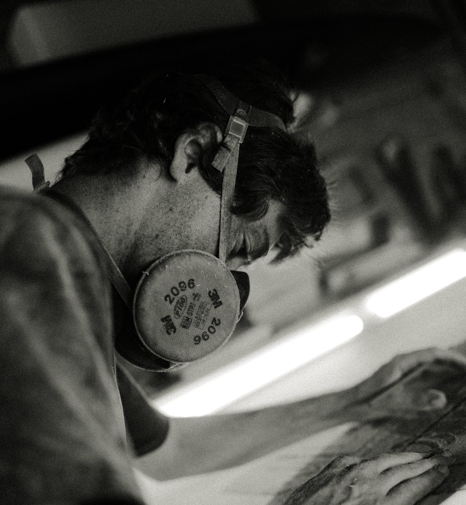 Black-and-white close-up of a masked surfboard builder at work.