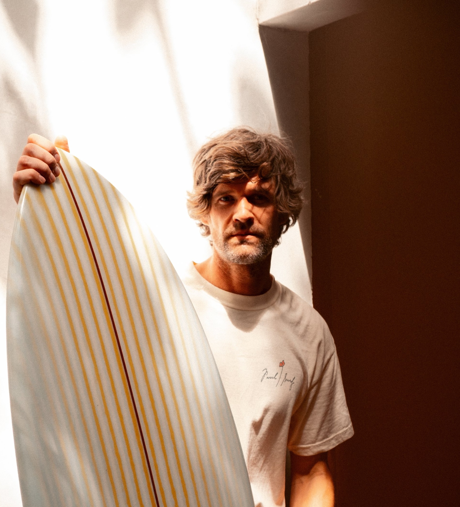 A board builder holds a white surfboard beside a bright window.