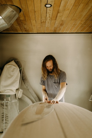 A shaper works on a surfboard beneath a wooden ceiling in the studio.