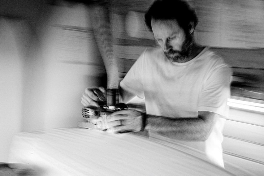 Black-and-white view of a shaper using a power tool on a surfboard.
