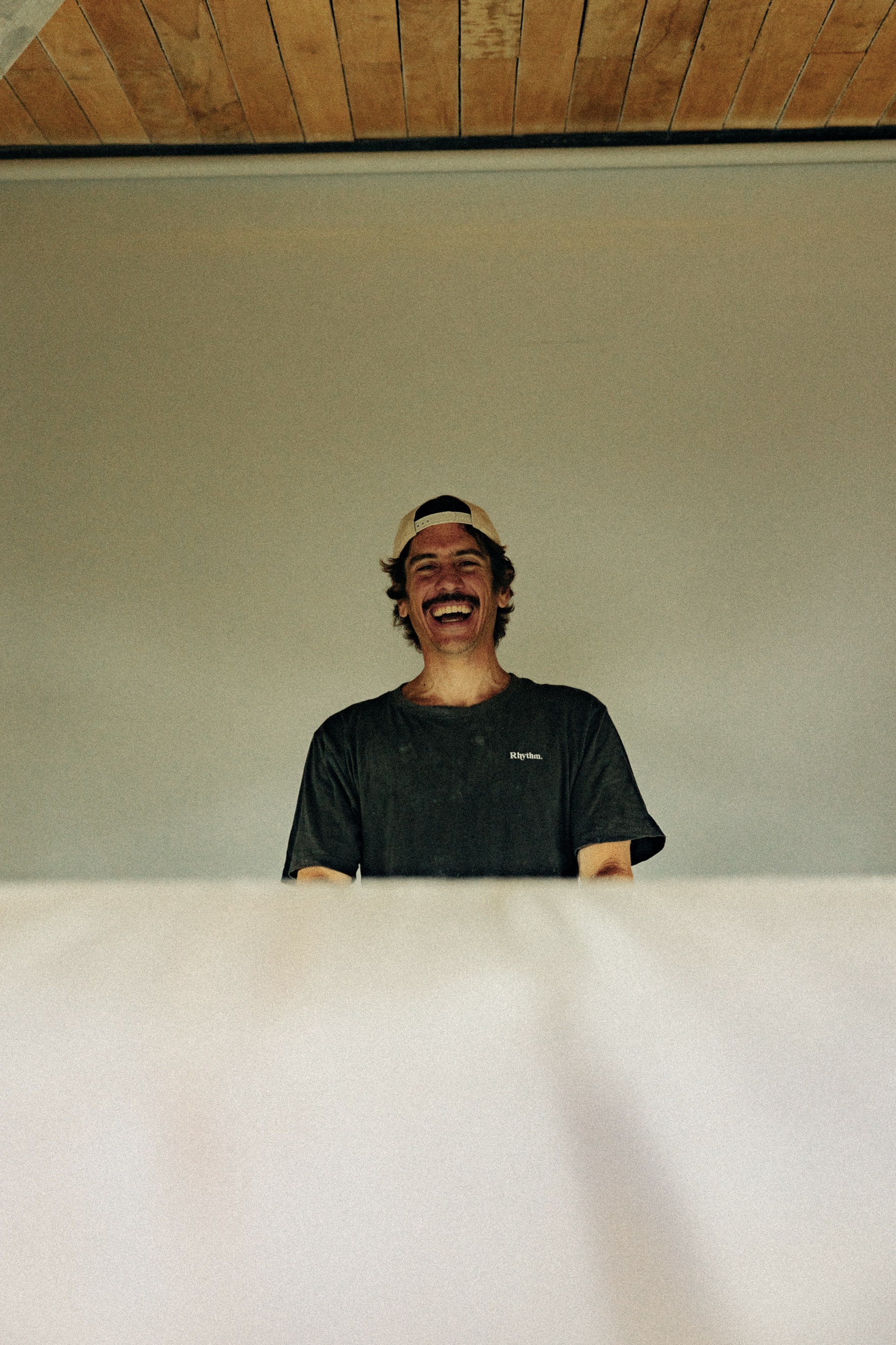 A shaper stands behind a white surfboard in a workshop.