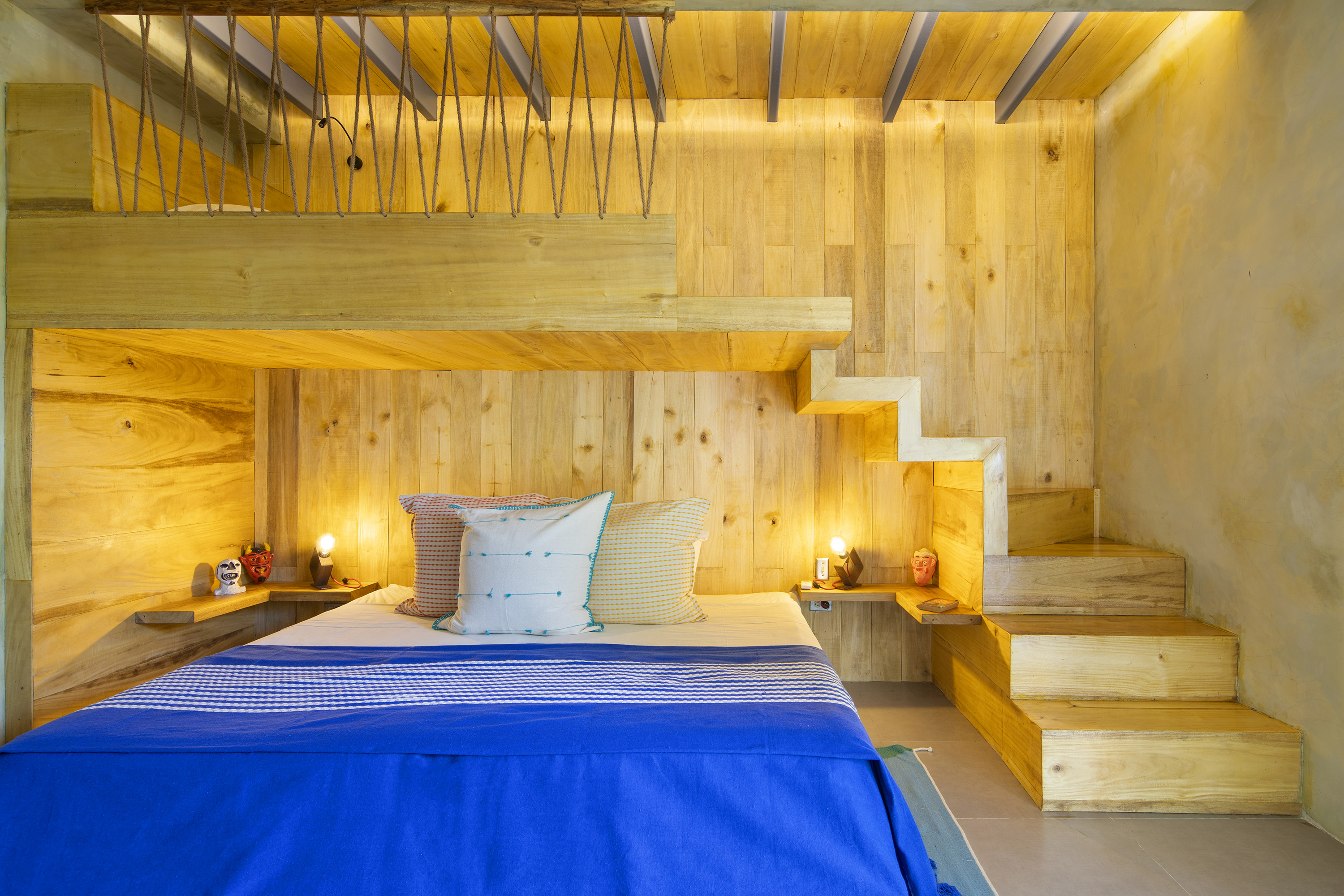 Cozy bedroom with light wood paneling on walls and ceiling, featuring a bed with blue blanket and three pillows, built-in wooden stairs to the right, two small shelves with lit lamps and decorative masks on each side of the bed.