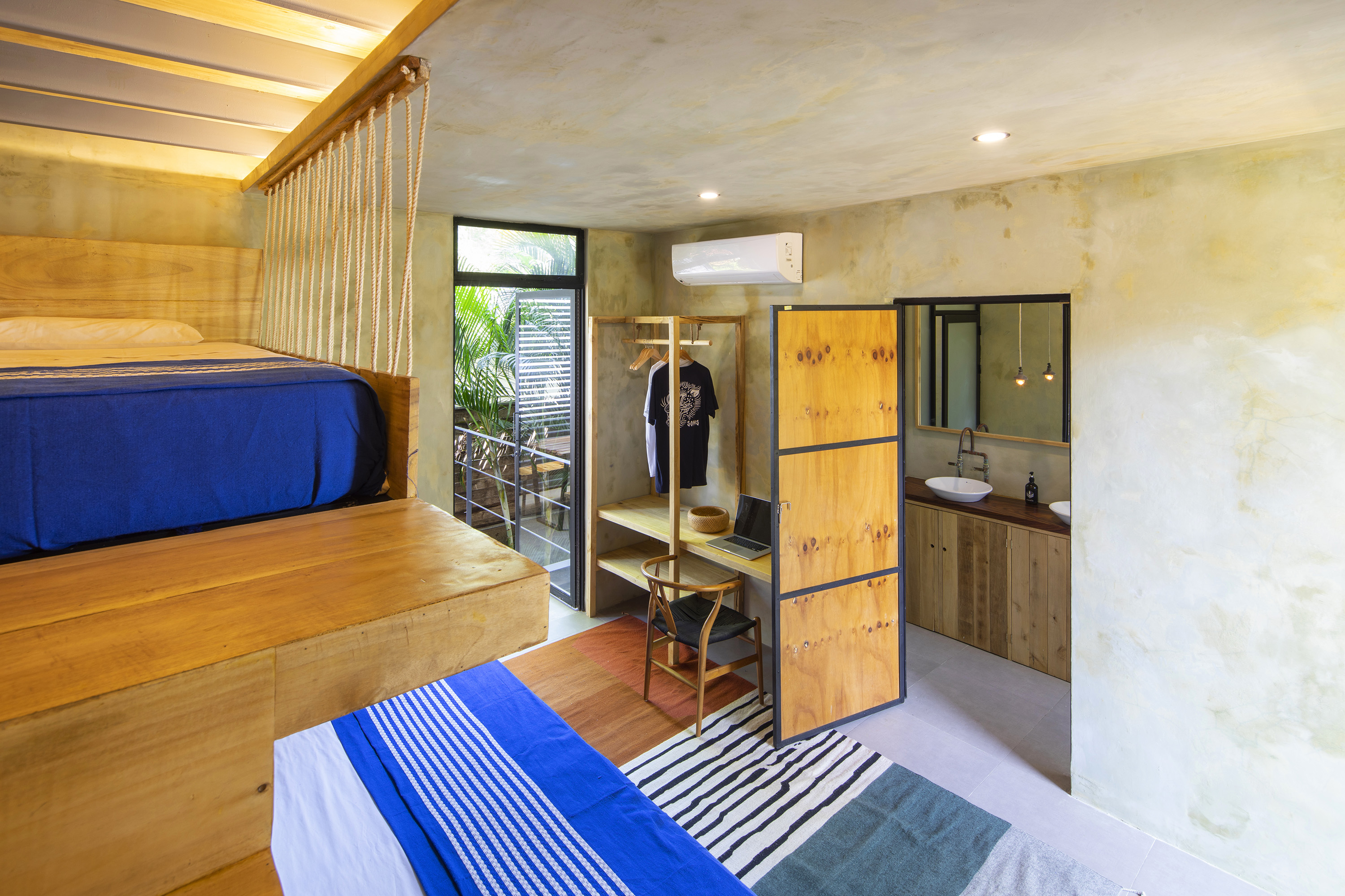 Modern bedroom with wooden loft bed and blue bedding, a striped rug, and a small desk with a chair, laptop, and hanging clothes near a glass door leading outside, next to a bathroom with a wooden vanity and two vessel sinks.