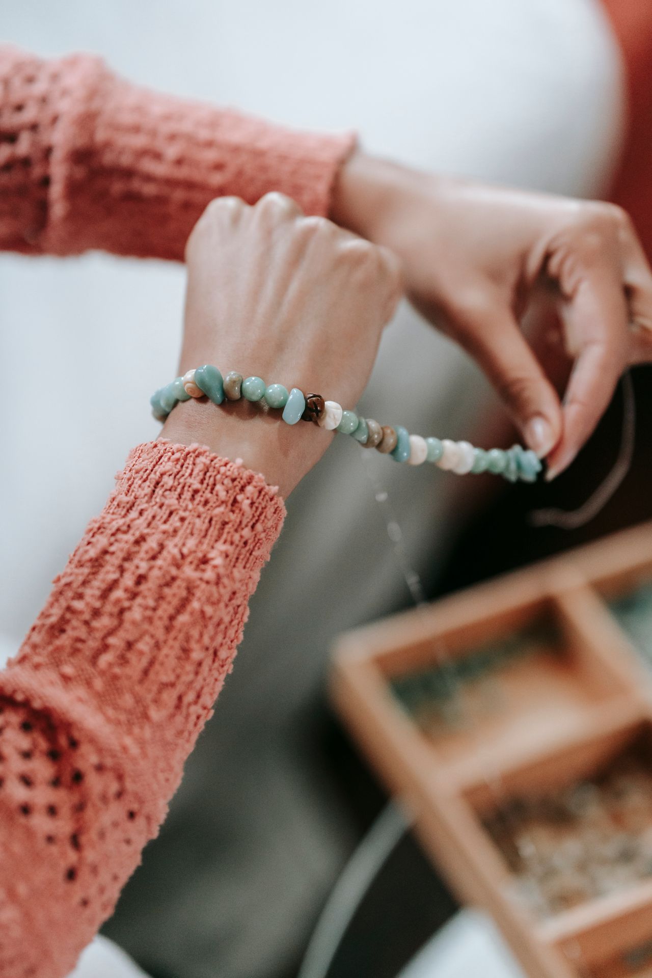 Hands of a person wearing a textured pink sweater stringing a beaded bracelet with green, blue, and white beads over a blurred wooden bead organizer.