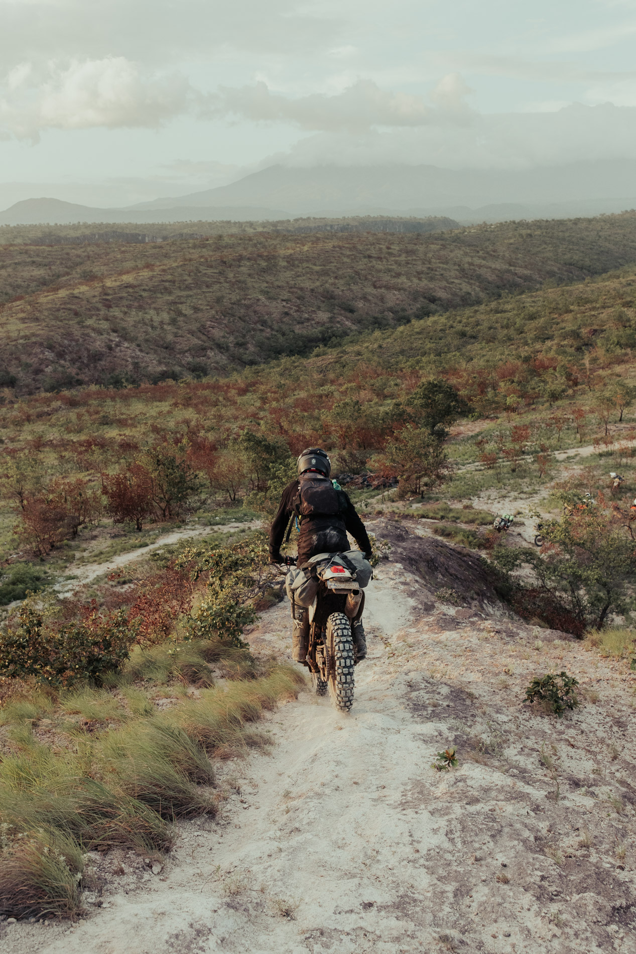 A person wearing a helmet and black clothing rides a dirt bike down a narrow, dusty trail on a hillside with green and brown vegetation and distant mountains under a cloudy sky.