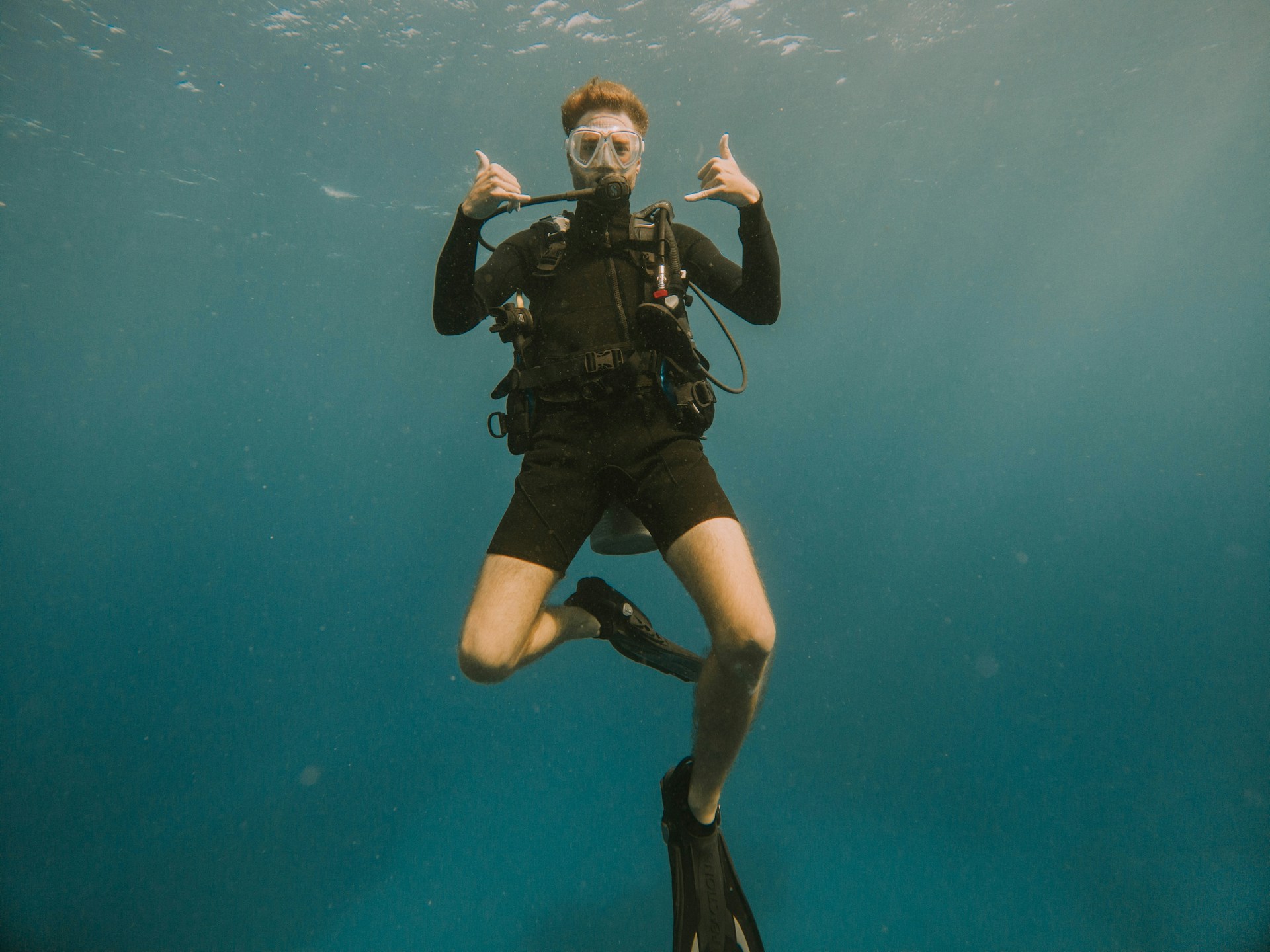 Scuba diver underwater wearing black wetsuit and fins, making a shaka hand gesture with both hands.