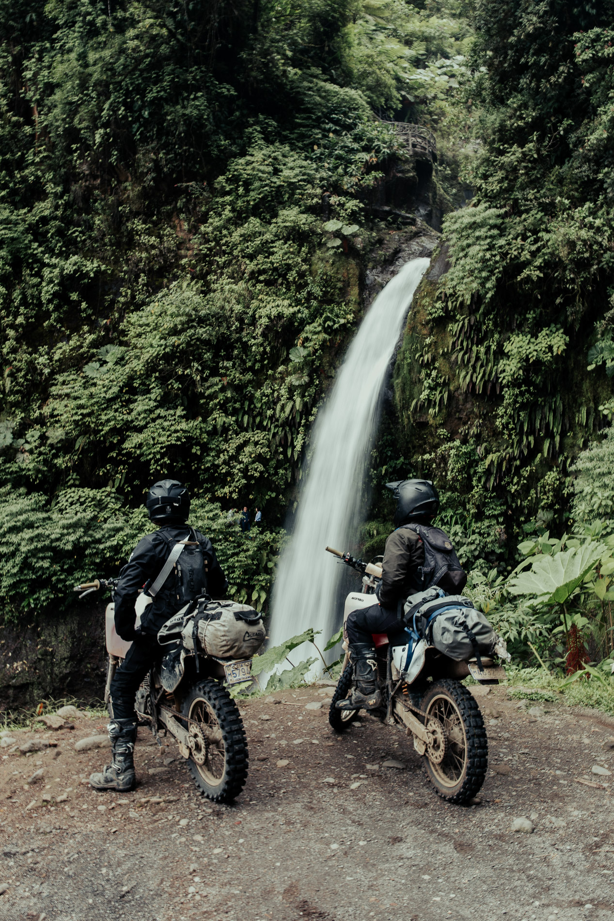 Two motorcyclists in black gear and helmets on dirt bikes with packed gear facing a tall, narrow waterfall surrounded by lush green foliage.