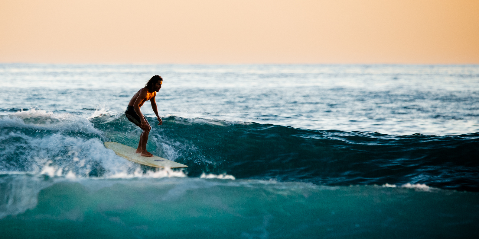 A man with long hair surfing on a wave in the ocean during sunset.
