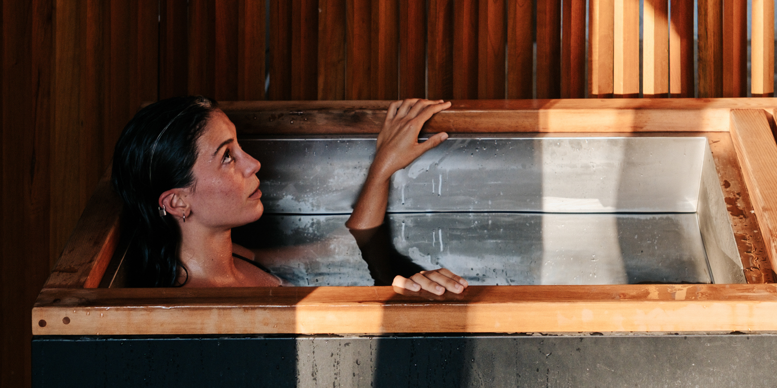 A woman with wet hair is sitting in a rectangular cold plunge pool with wooden and metal edges inside a wooden-paneled room, looking upwards with her hands resting on the pool's edge.
