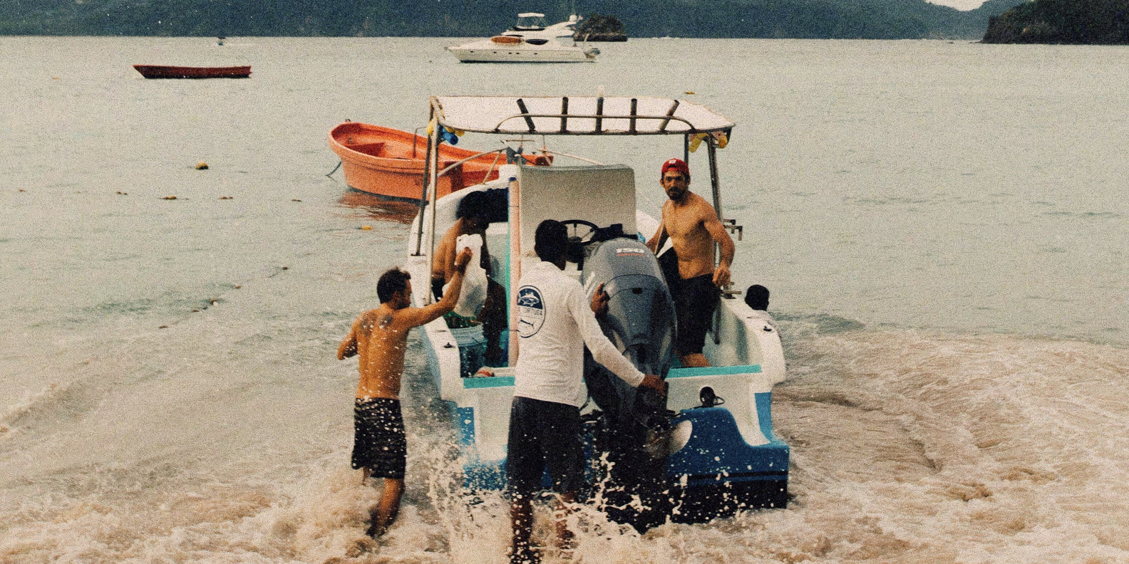 Four men around a small motorboat in shallow water near the shore, two men standing in the water pushing the boat while two men stand or sit in the boat with one wearing a red cap.