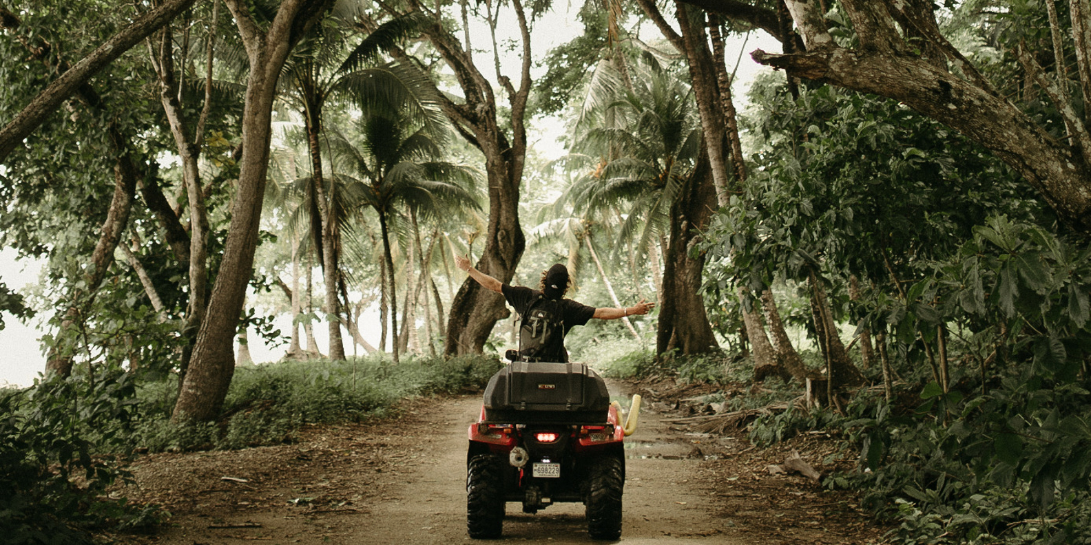 Person with a backpack riding a red ATV on a forest trail surrounded by tall trees and dense green foliage, raising both arms outward.