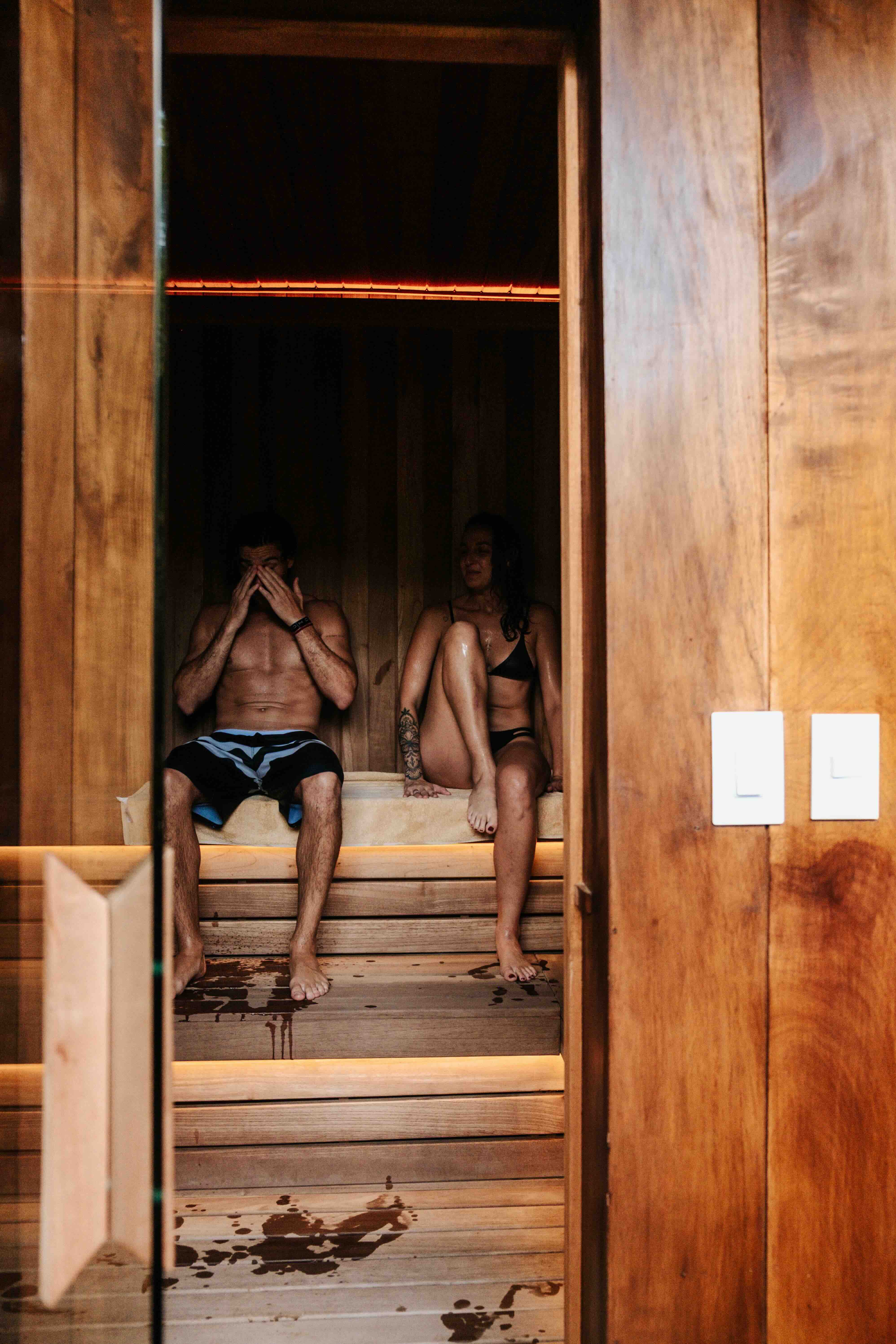 A man and a woman wearing swimwear sit inside a wooden sauna with glass doors; the man covers his face with his hands while the woman looks at him, with water droplets on the floor.