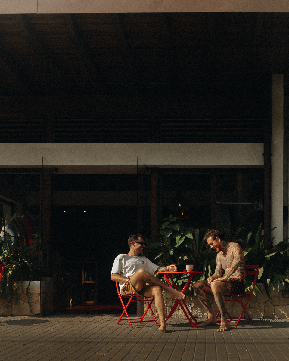 Two men sitting barefoot on red folding chairs outside a building, chatting and laughing with plants in the background and a red table between them holding a coffee cup.