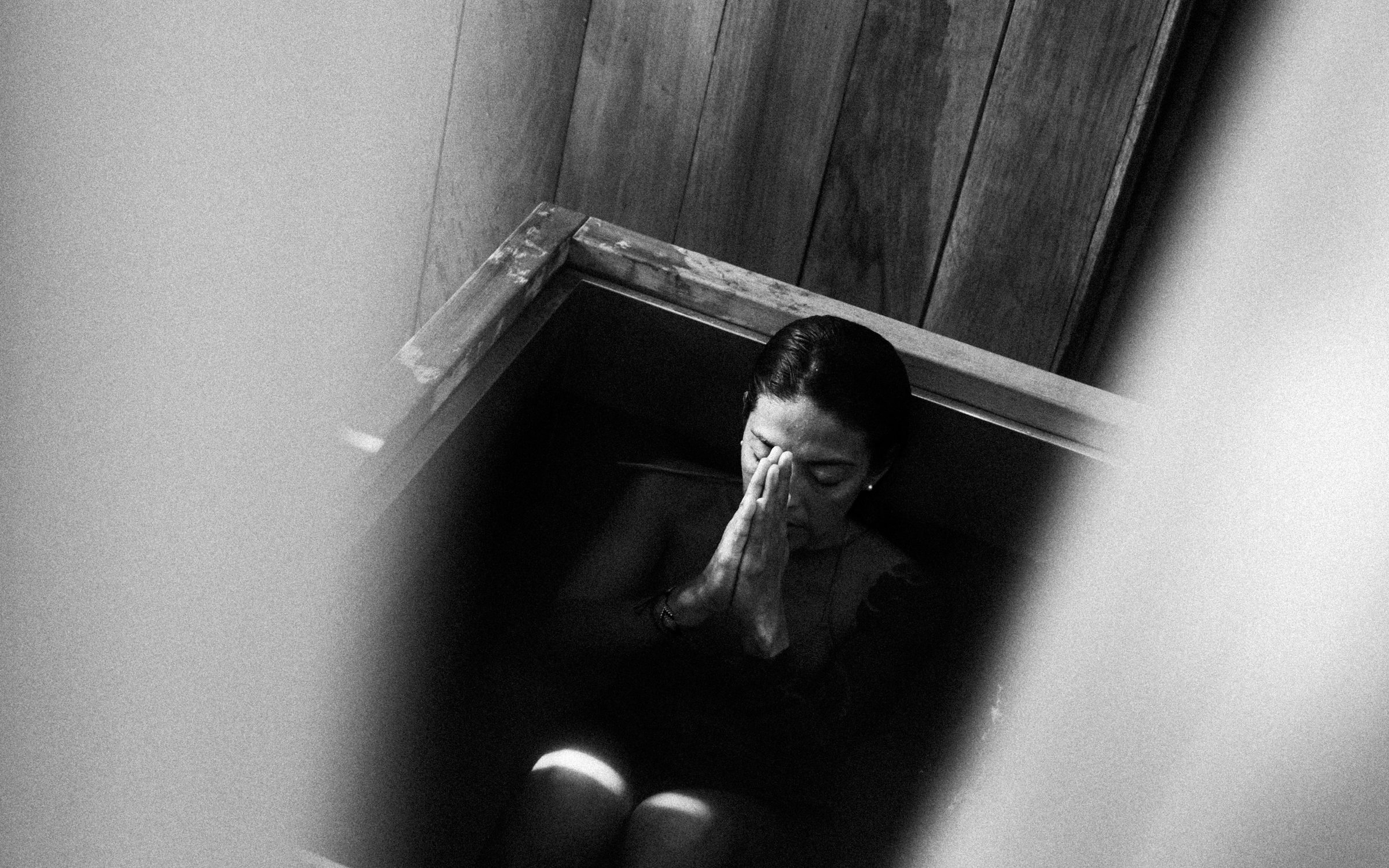 A woman with closed eyes and hands pressed together in a prayer or meditation pose, sitting inside a wooden bathtub filled with dark water, captured in black and white.