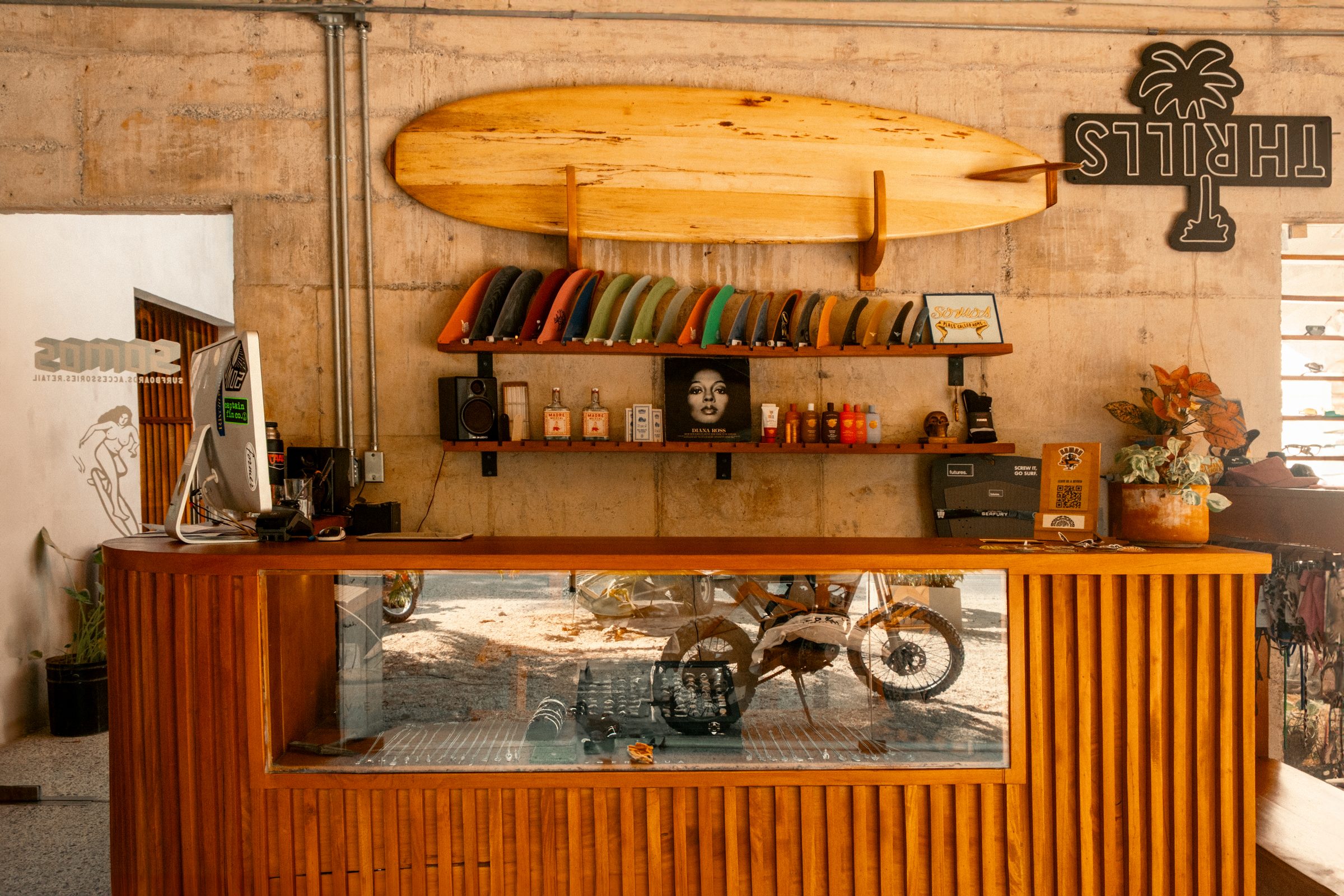 Wooden shop counter with glass display case showing accessories and jewelry, with a row of surfboard fins on wall-mounted shelves above and a wooden surfboard mounted on the wall, alongside bottles of sunscreen and products, a plant in a pot, and a computer monitor on the counter.