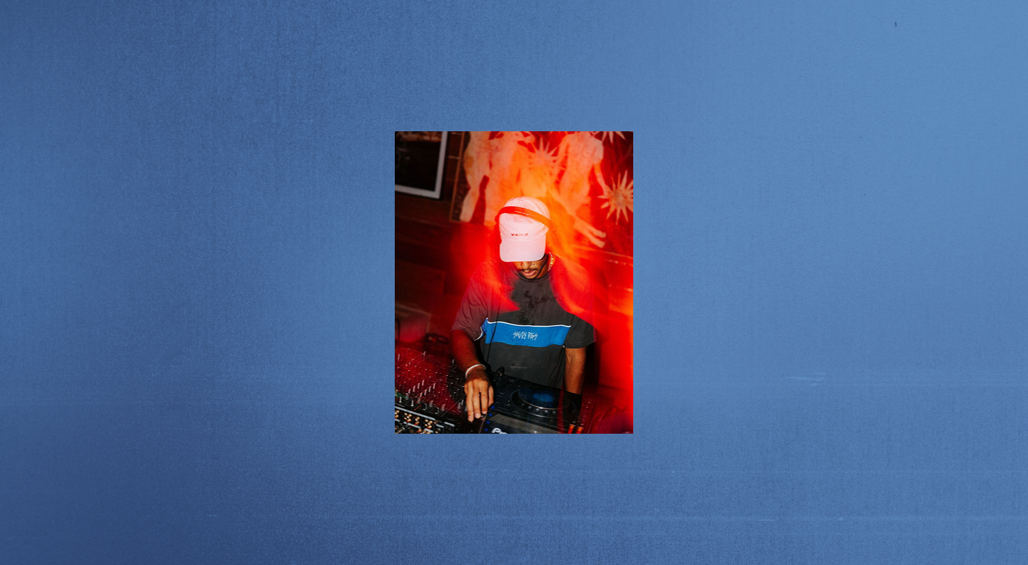 DJ wearing a white cap and headphones mixing music on a turntable with red lighting effects and artistic wall art in the background.