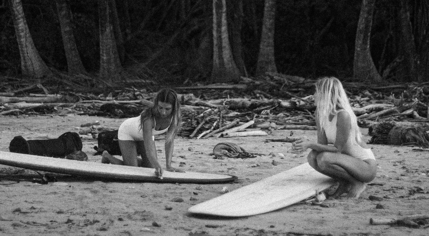 Two women in swimsuits on a sandy beach preparing white surfboards surrounded by driftwood and trees, one kneeling and the other crouching.