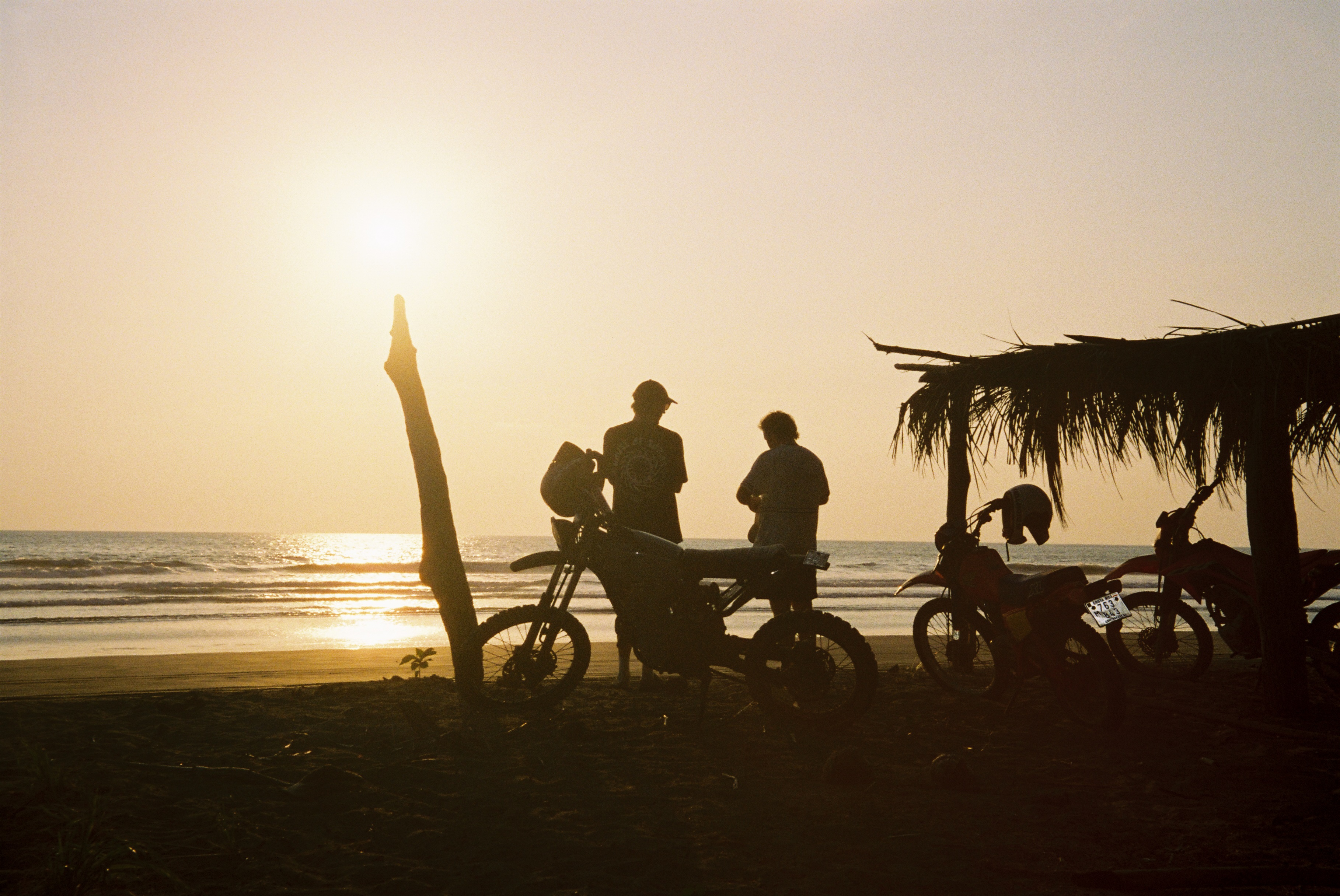 Silhouettes of two people and three motorcycles on a beach at sunset, with the sun low over the ocean and a small thatched shelter on the right.