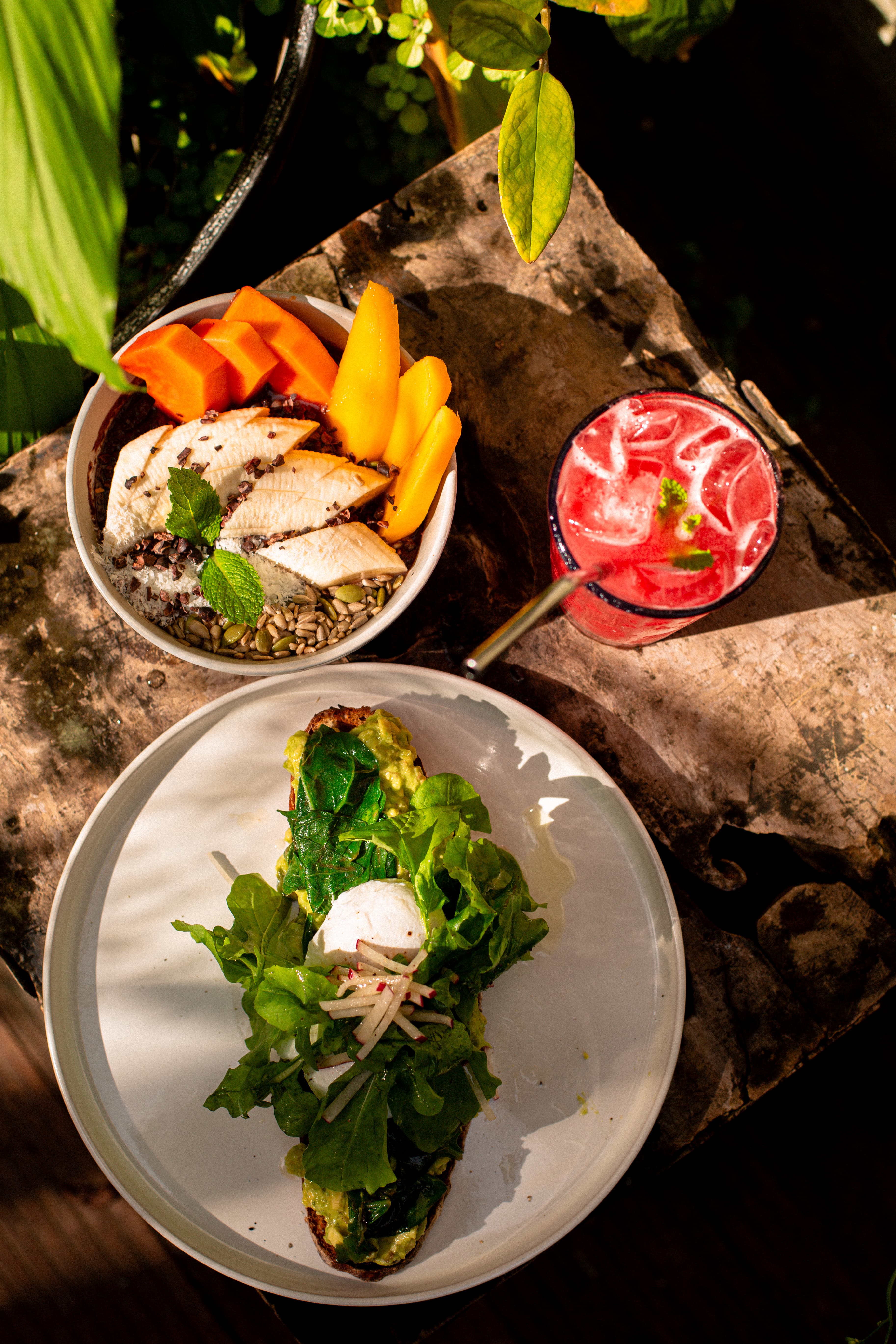 Top-down view of a breakfast spread on a rustic wooden surface including a bowl with sliced banana, papaya, mango, seeds, and mint leaves, a glass of pink iced drink with a straw and mint garnish, and a plate with avocado toast topped with leafy greens, a poached egg, and sliced radish.