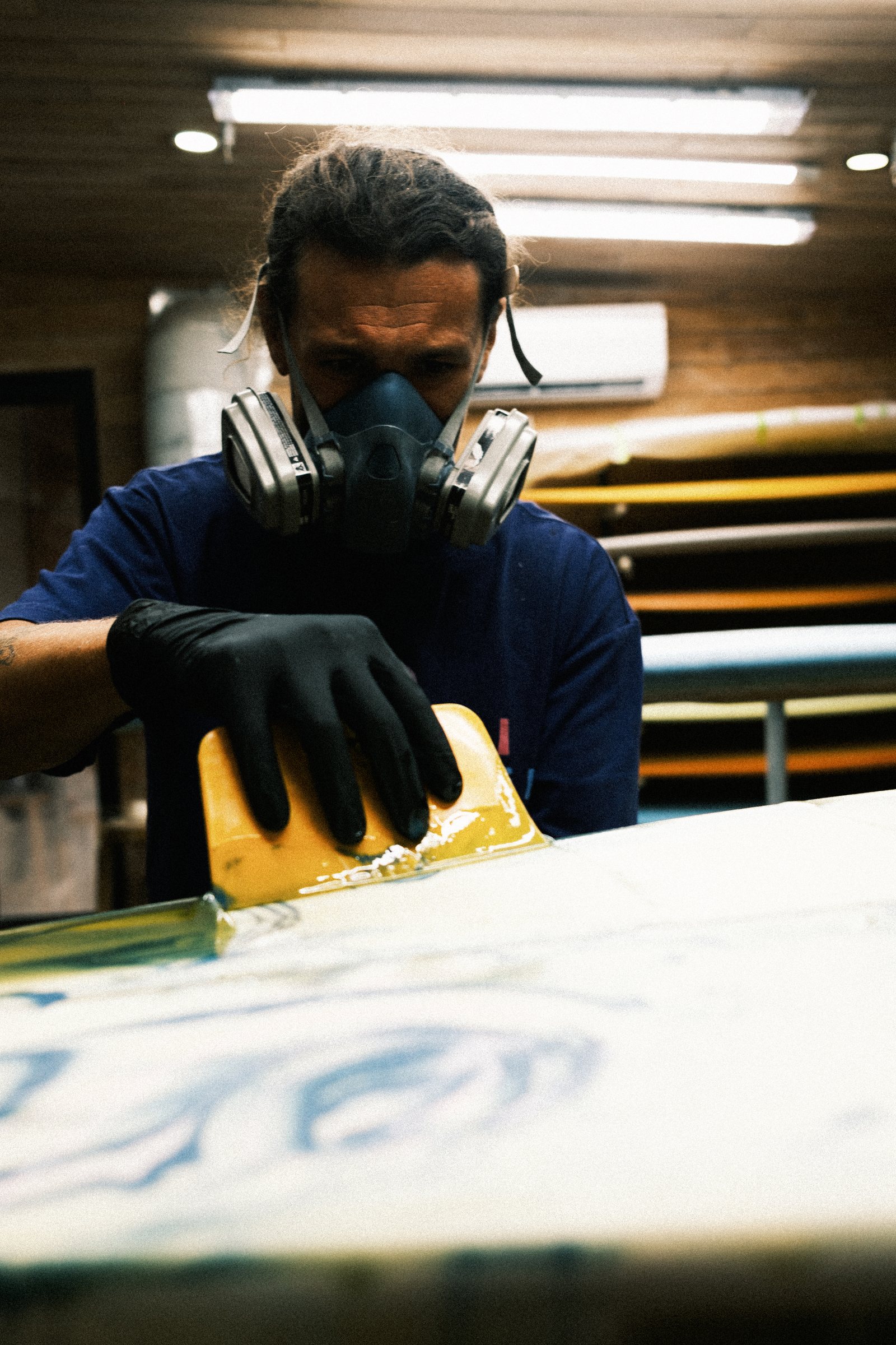 Man wearing a respirator mask and black gloves working on a surfboard in a workshop.