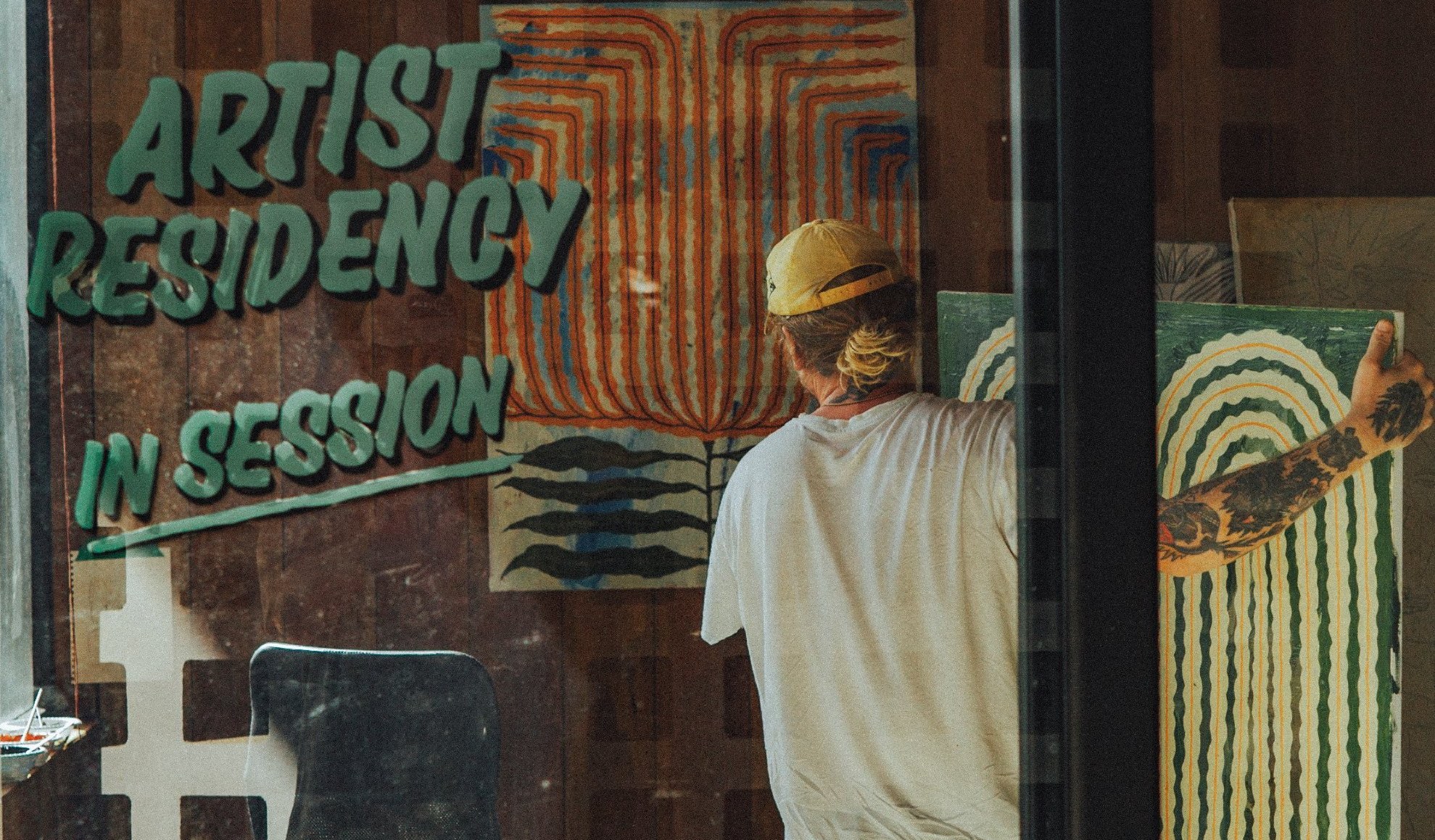 An artist in a yellow cap hangs a green striped painting in the House of Somos studio, behind a window painted with Artist Residency In Session