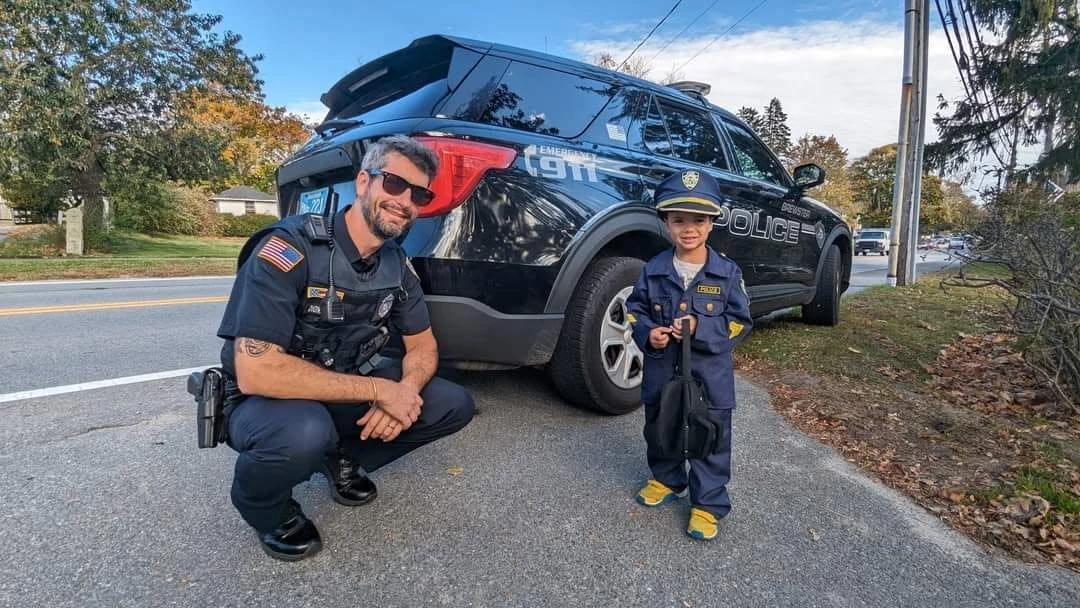 A Brewster officer kneels beside a smiling child dressed in a police costume in front of a patrol SUV