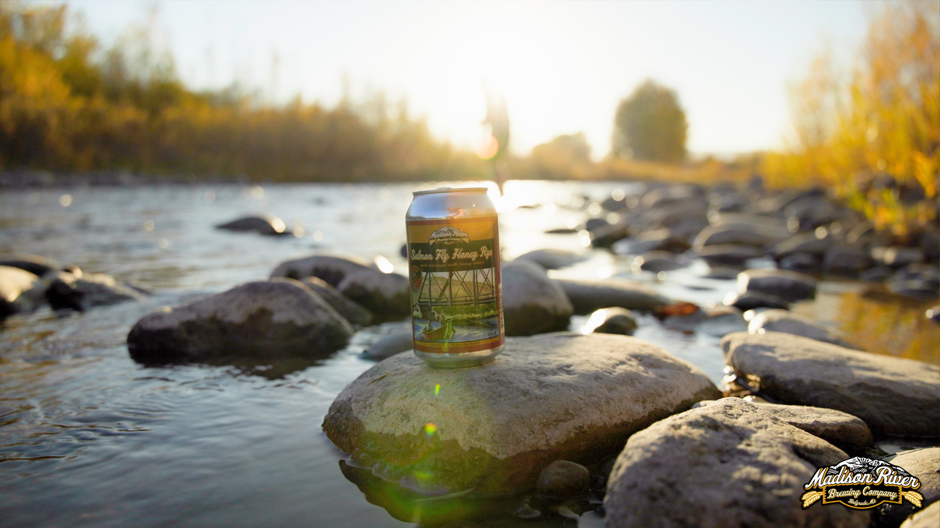 A can of Madison River Brewing Company's Salmon Fly Honey Rye beer placed on a rock in a shallow river with many stones and the setting sun in the background.