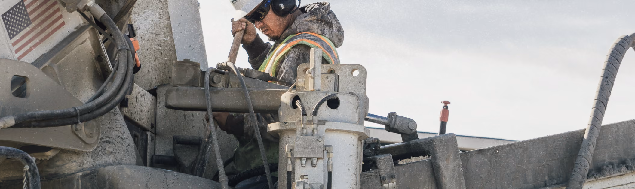 Worker wearing safety glasses and ear protection operating industrial machinery outdoors under a cloudy sky.