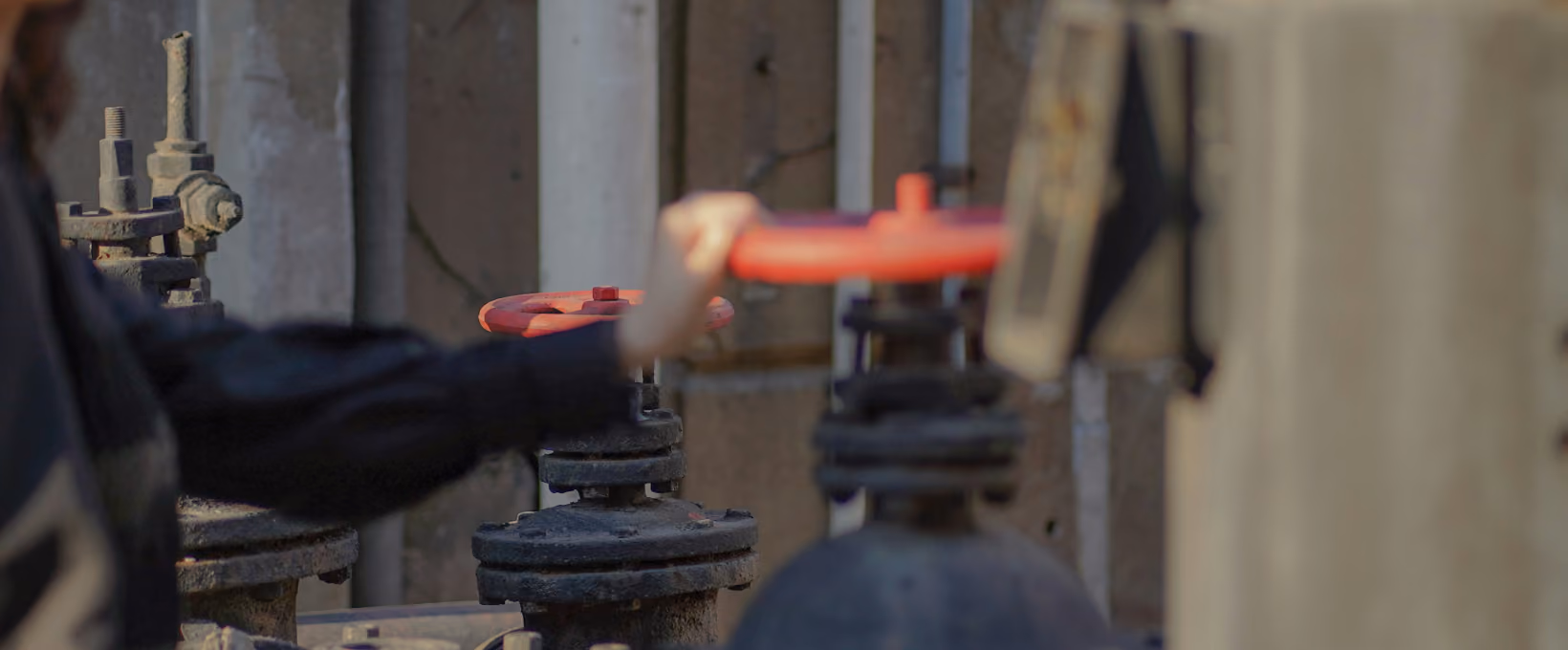 Person's hand turning a red valve wheel on industrial pipes in a dimly lit facility.