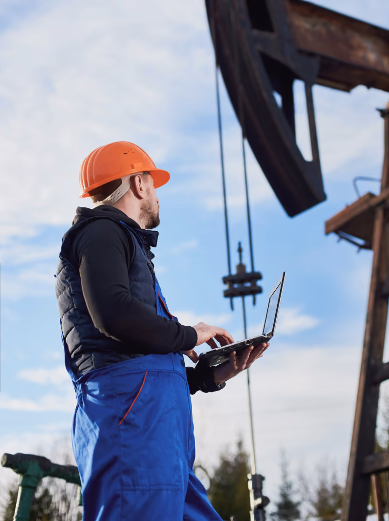 Worker wearing an orange hard hat and blue overalls using a laptop outdoors near industrial machinery against a partly cloudy sky.