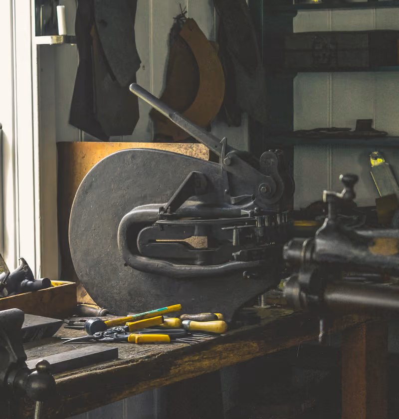 A vintage metal press machine on a wooden workbench in a dimly lit workshop, surrounded by various hand tools with yellow and green handles.