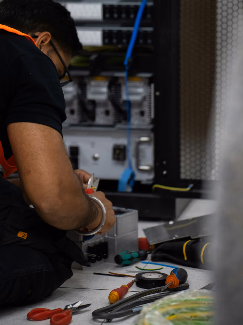 Man wearing glasses and a black shirt working on electrical components with tools like screwdrivers, electrical tape, and wire cutters on the floor near a server rack.