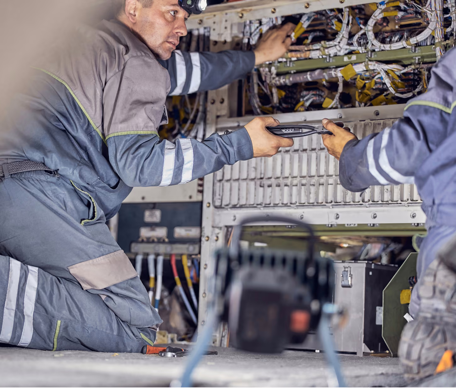 Two technicians in reflective workwear working on complex wiring inside an industrial or aviation panel, one handing a tool to the other.
