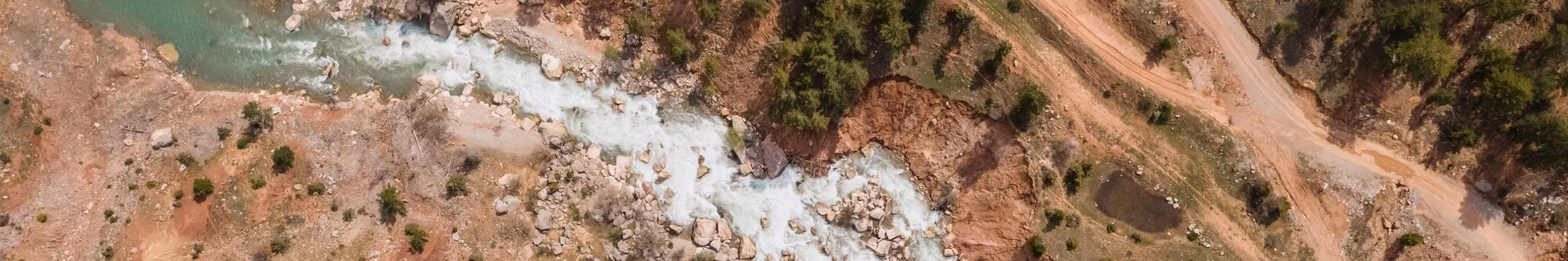 Aerial view of a winding white river flowing through a rocky landscape with sparse greenery and dirt paths on both sides.
