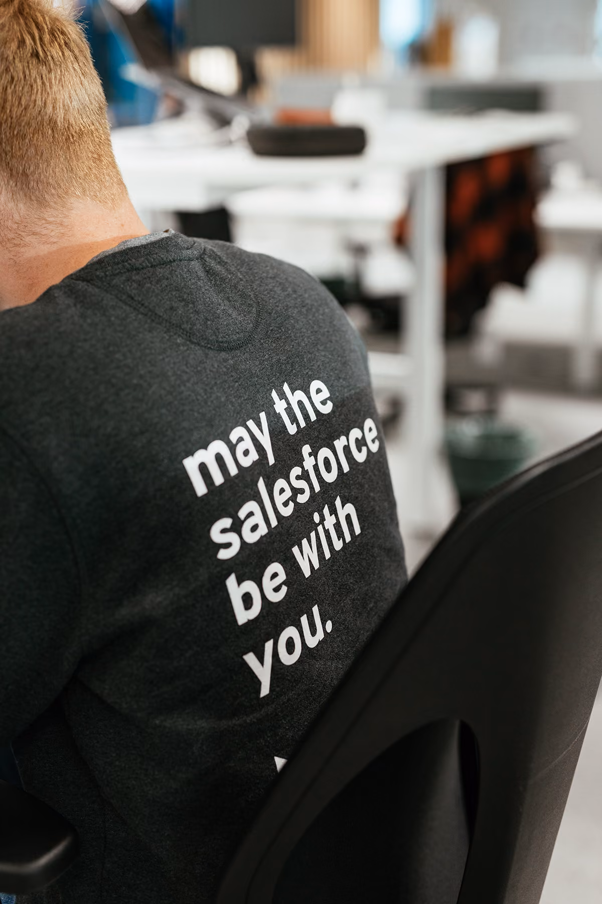 Person with short blonde hair sitting on a black chair wearing a dark gray shirt with white text that reads 'may the salesforce be with you' in an office environment.