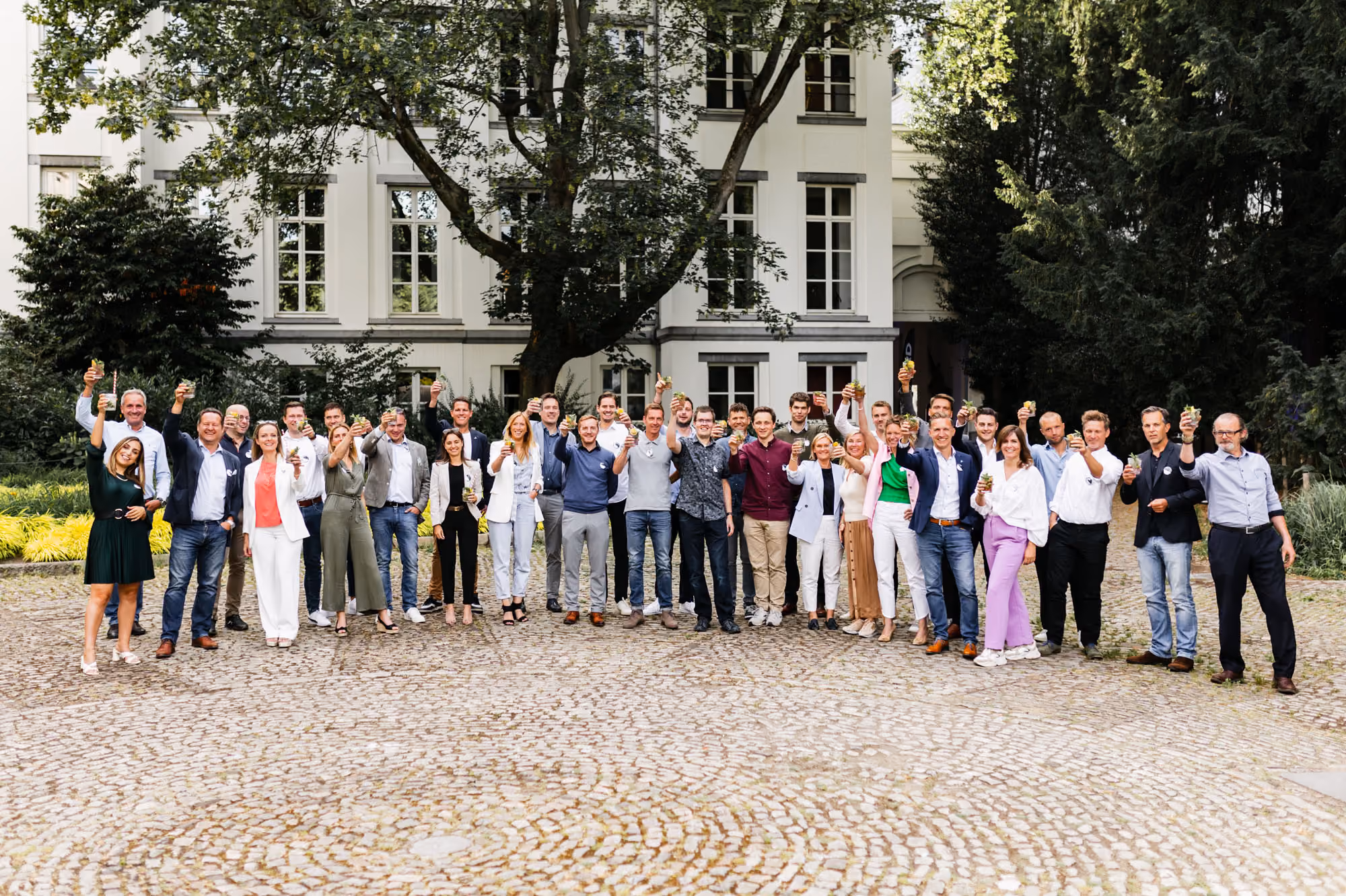 Large group of men and women standing outdoors on a paved area in front of a white multi-story building with trees, all raising drinks and smiling at the camera.