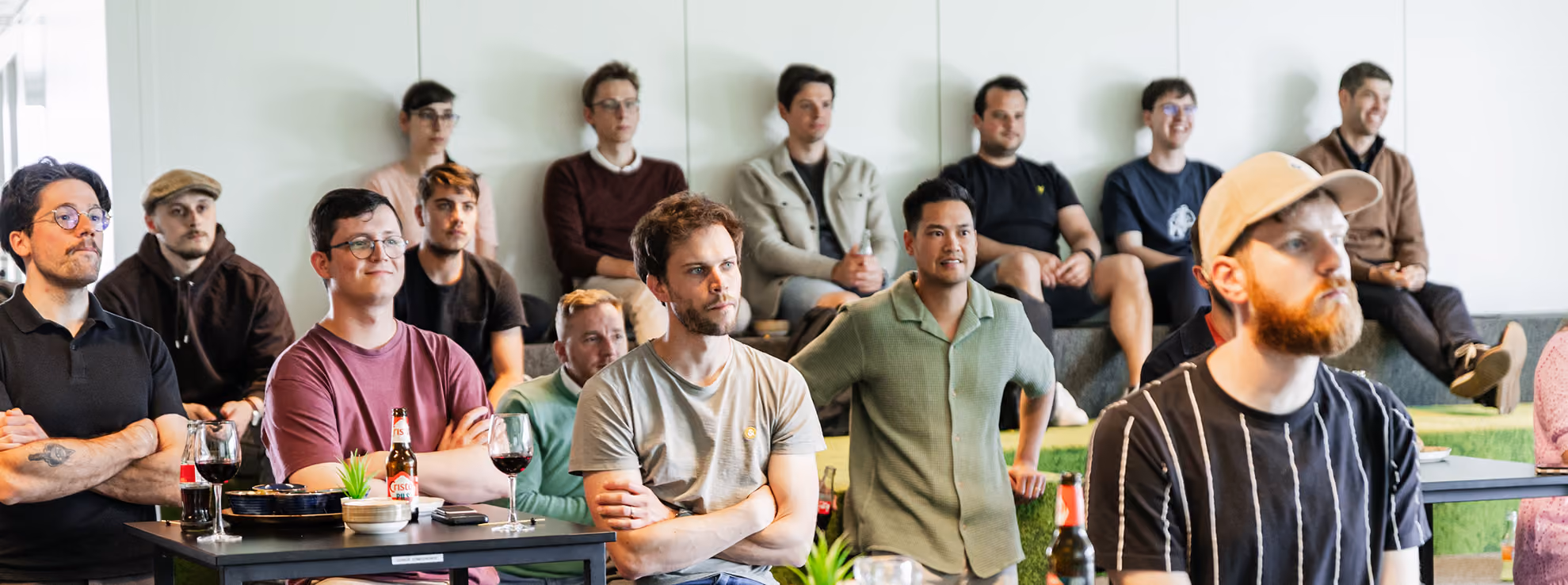 Group of diverse men sitting attentively in a modern room, some with drinks on tables, likely attending a presentation or meeting.