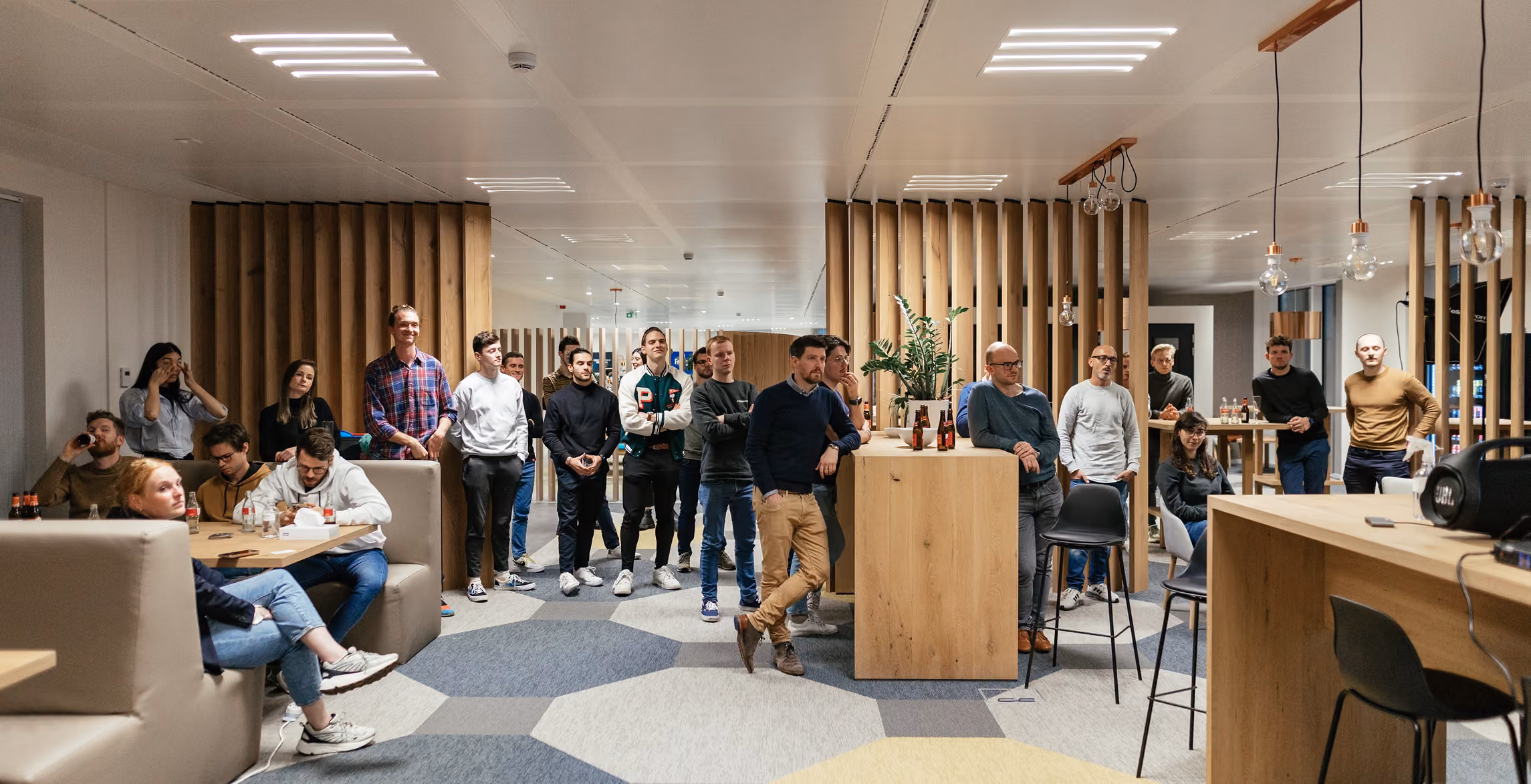 Group of people standing and sitting in a modern office lounge area with wooden paneling, geometric carpet, and hanging light bulbs, casually socializing and listening to something outside the frame.
