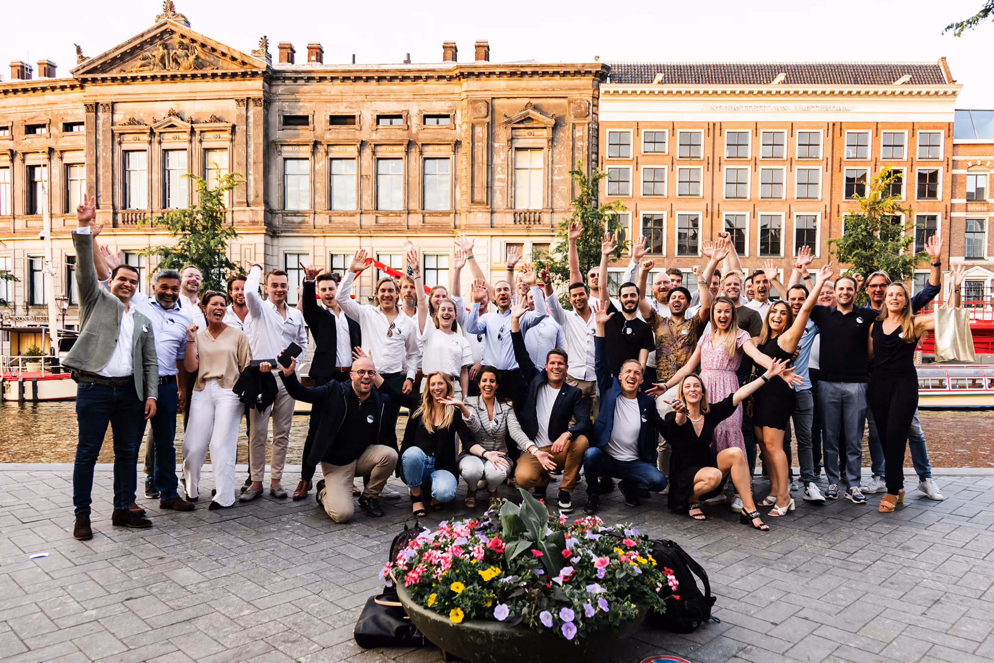 A large group of about 30 people outdoors on a paved area near a body of water, posing happily with raised hands, in front of historic buildings during daylight.