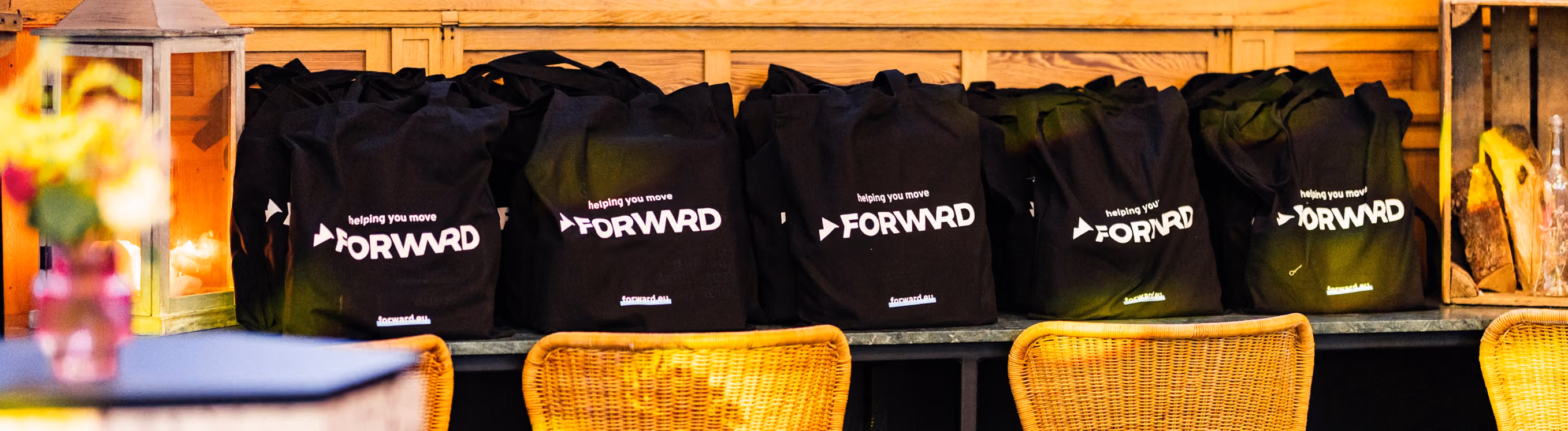 Row of black tote bags with the text 'helping you move FORWRD' lined up on a bench behind woven chairs in a warmly lit setting.