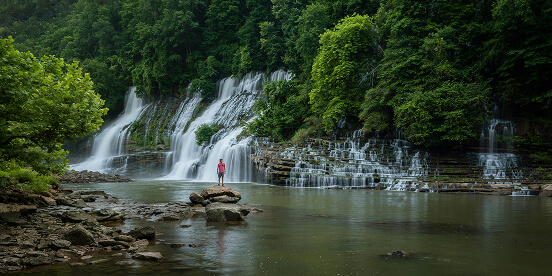 East Tennessee Attractions: Twin waterfalls pouring down a rock face into a wide pool, with one person standing on a rock for scale.