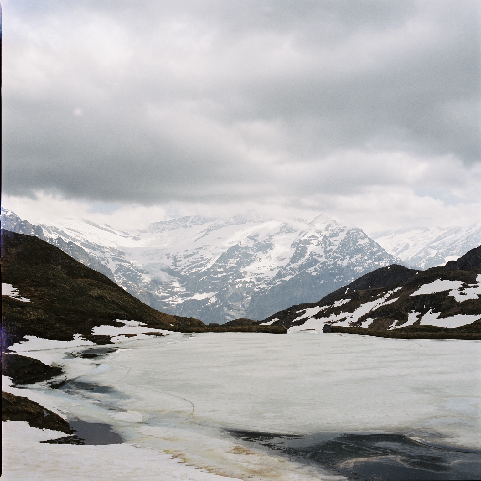 Snow-covered mountains beneath a cloudy sky, reflected in a partially frozen alpine lake.
