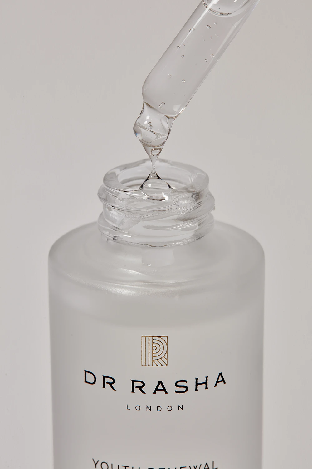 Dropper releasing clear serum into an open frosted glass bottle labeled Dr Rasha London.