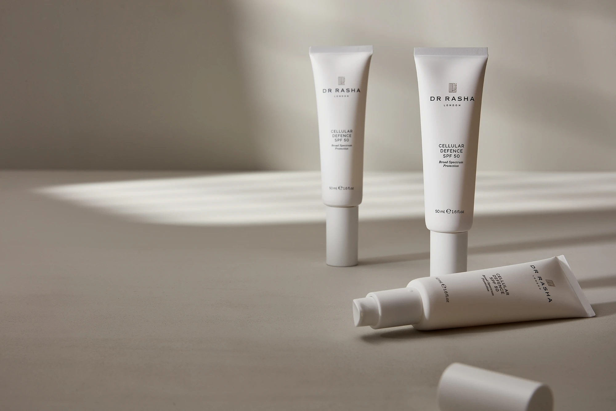Three Dr Rasha Cellular Defence SPF 50 skincare tubes on a beige surface with soft striped light.