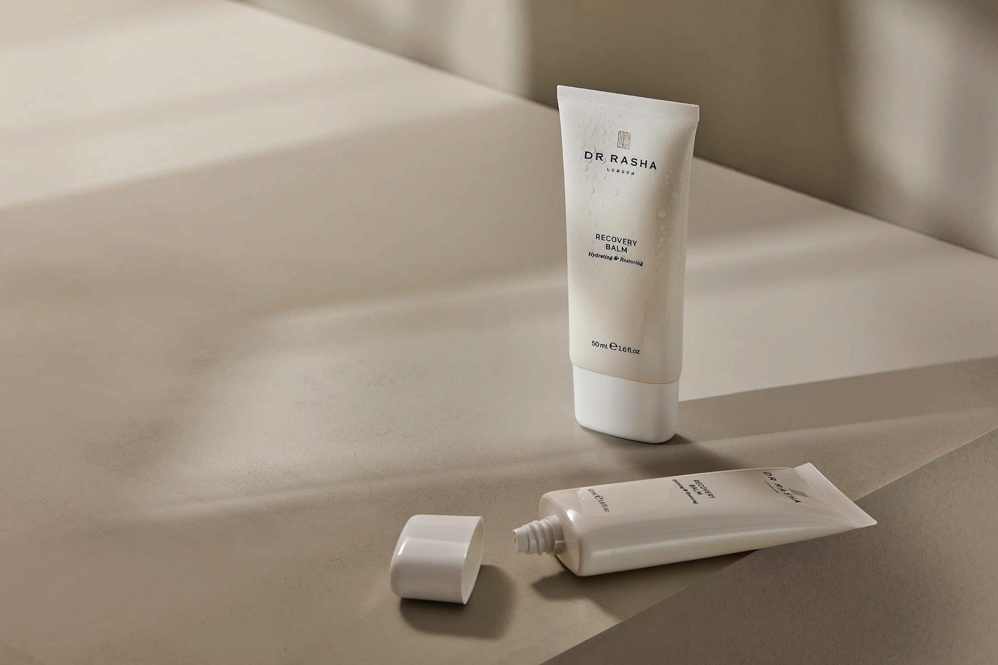 Two white tubes of Dr Rasha Recovery Balm on a beige surface, one standing and one lying with cap off.