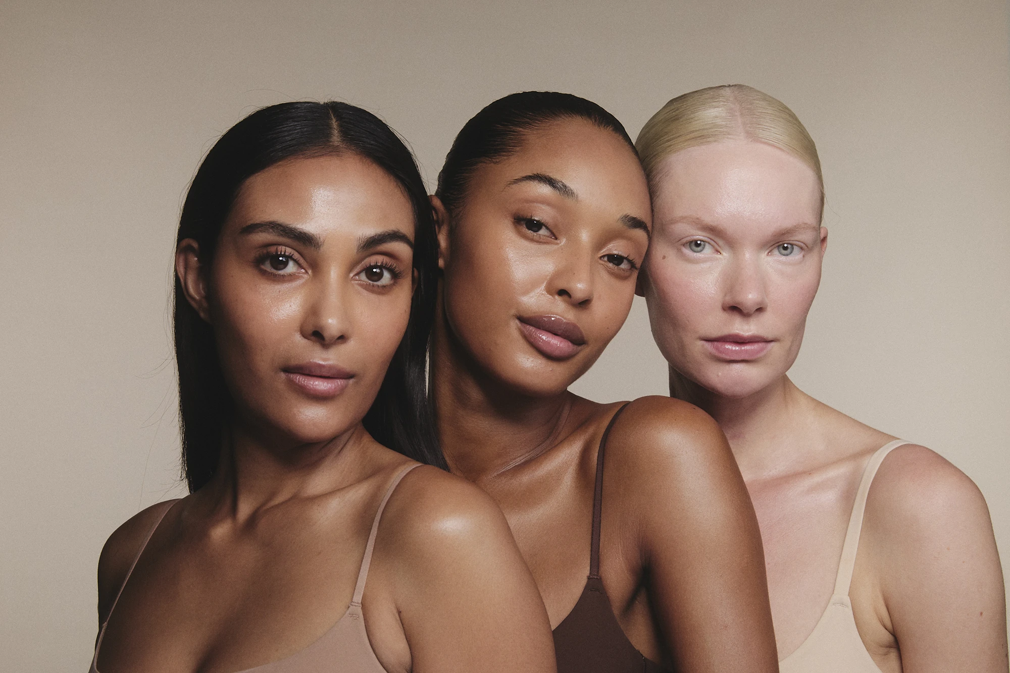 Three women of diverse skin tones posing closely together against a plain background.
