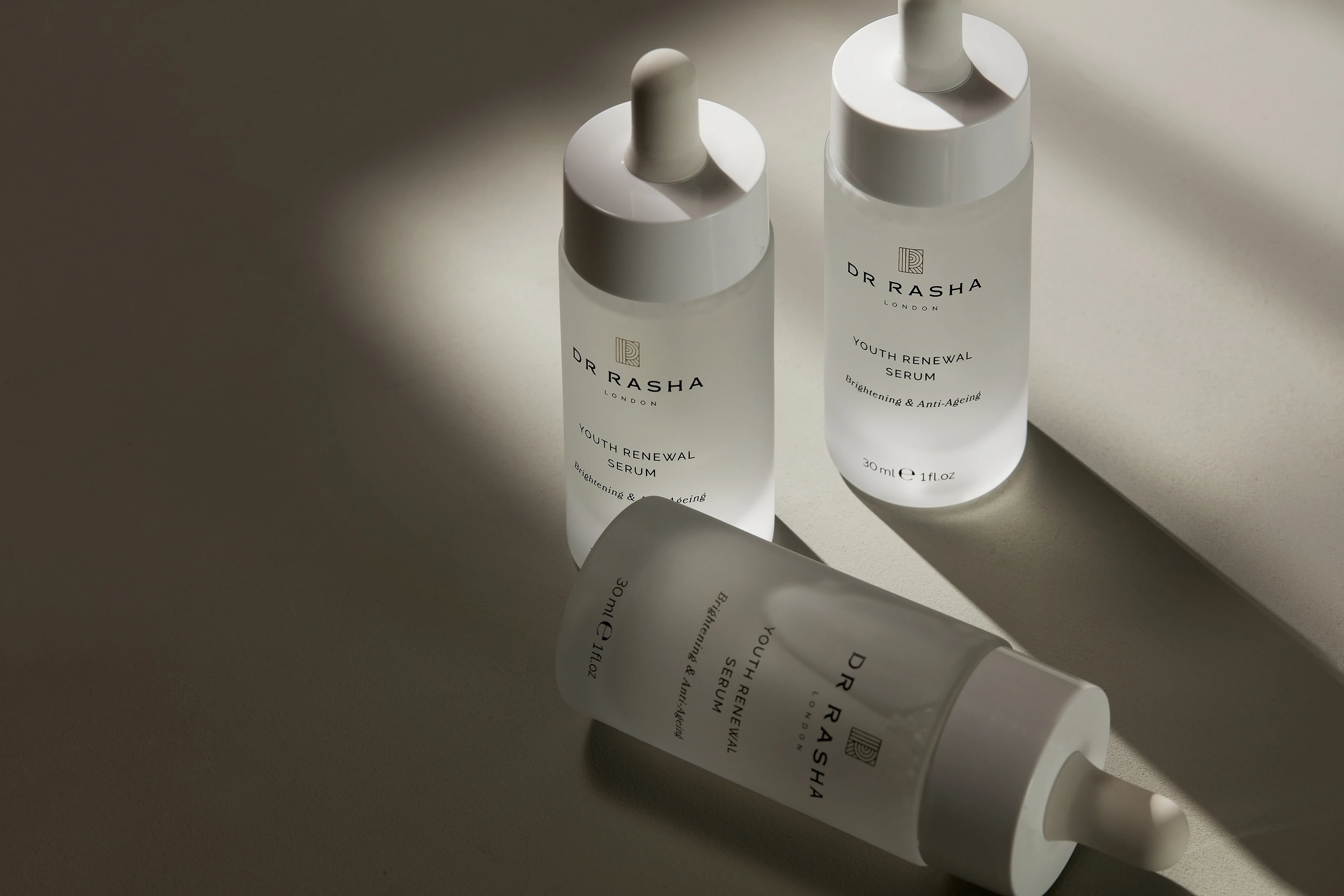Three Dr Rasha Youth Renewal Serum dropper bottles casting shadows on a beige surface.